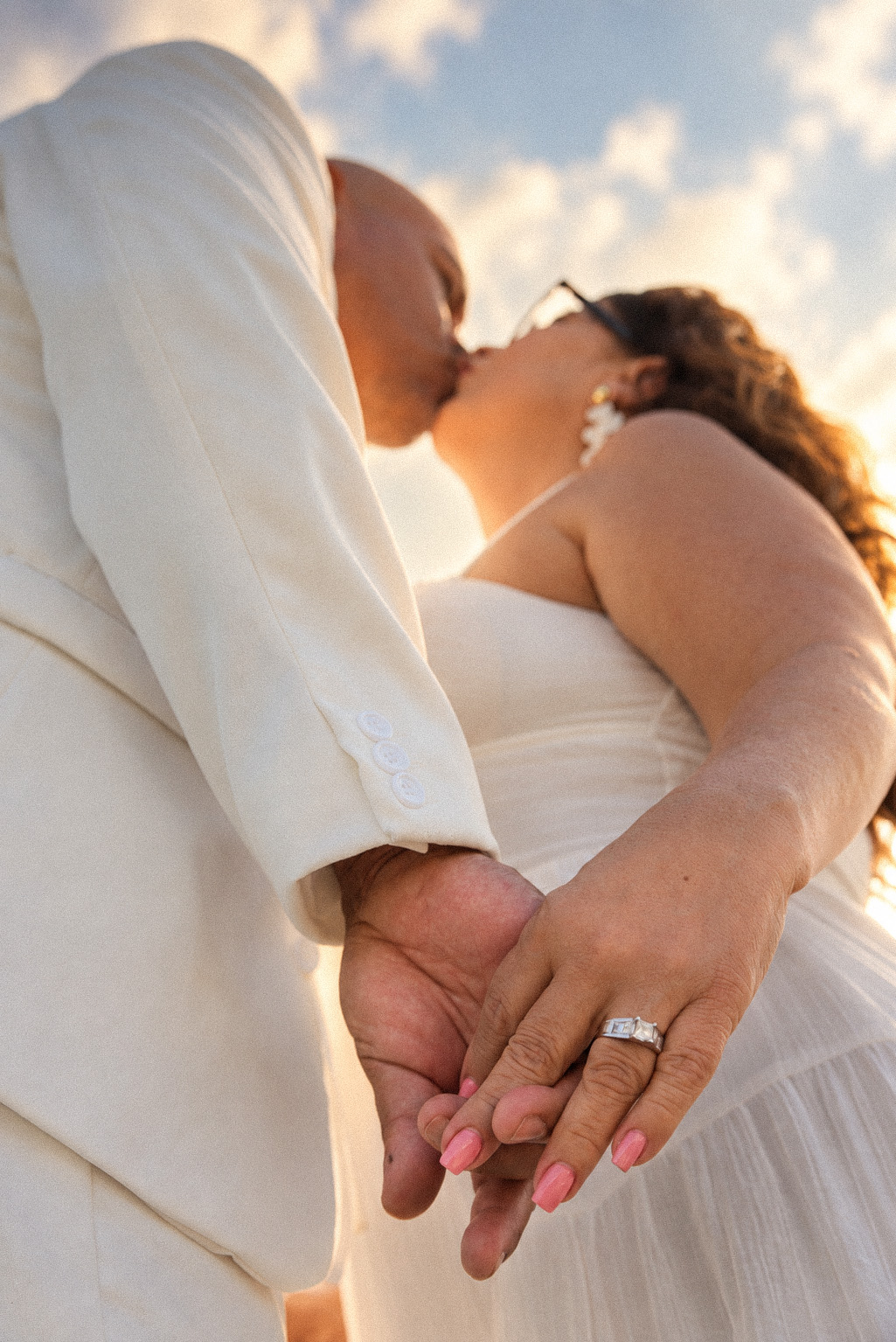 Couple in white sharing a kiss against a golden sky, joined hands in the foreground